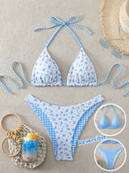A two-piece bikini set laid flat on a light beige textured background, featuring a white base with blue floral print on one side and blue gingham check on the other. The set includes a halter triangle bikini top and matching low-rise bikini bottoms.