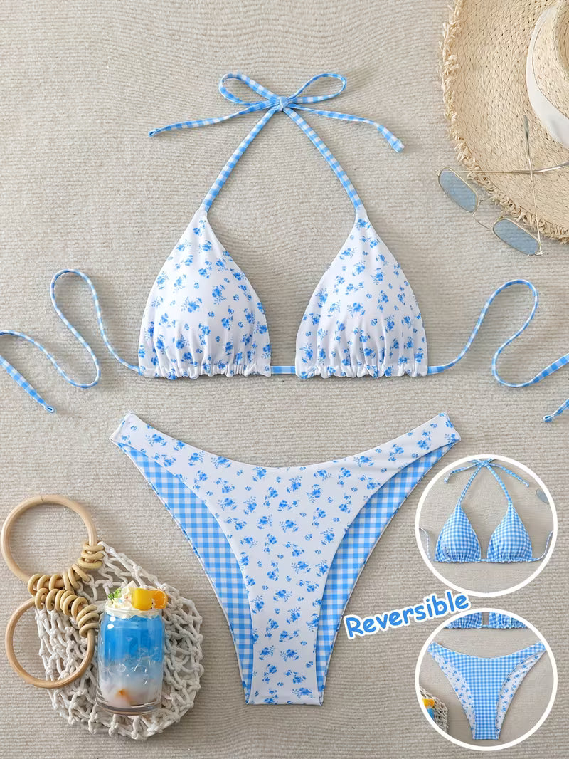 A two-piece bikini set laid flat on a light beige textured background, featuring a white base with blue floral print on one side and blue gingham check on the other. The set includes a halter triangle bikini top and matching low-rise bikini bottoms.