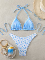 A two-piece bikini set laid flat on a beige textured surface, featuring a light blue and white gingham triangle top with tie straps and matching bottoms with a small blue floral print. A straw hat, sunglasses, and a small drink in a crochet bag are styled around the swimwear.