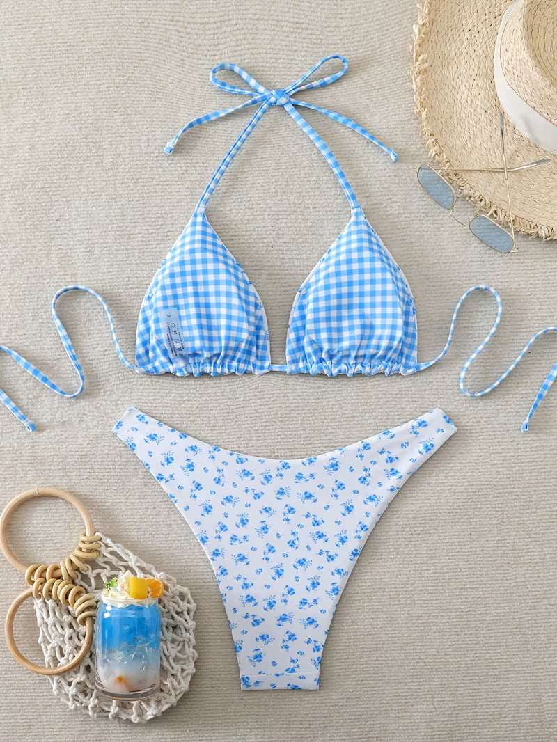 A two-piece bikini set laid flat on a beige textured surface, featuring a light blue and white gingham triangle top with tie straps and matching bottoms with a small blue floral print. A straw hat, sunglasses, and a small drink in a crochet bag are styled around the swimwear.