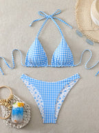 A two-piece string bikini laid flat on a beige textured surface. The bikini features a light blue and white gingham pattern on the top and a matching blue patterned print on the bottoms, with a white inner lining.