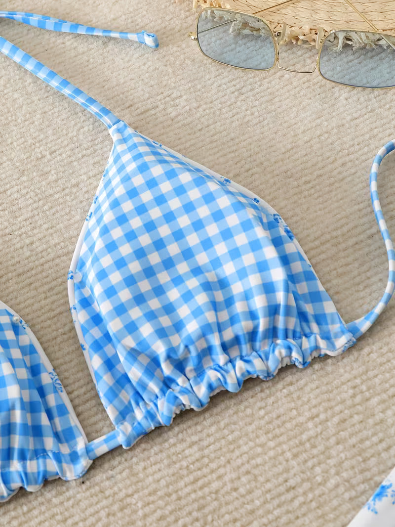 A close-up of a blue and white gingham bikini top laid flat on a beige textured surface, with part of a straw hat and sunglasses visible at the top edge.