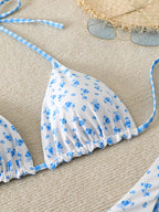 A close-up of a bikini top laid flat on a beige textured surface, featuring a white base with a blue floral print and light blue gingham trim. A straw hat and sunglasses are partially visible as styling props.