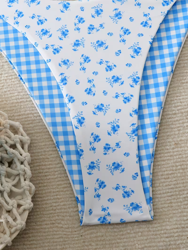 A close-up of a bikini bottom laid flat on a beige textured surface, featuring a white front panel with small blue floral print and a light blue gingham side panel.