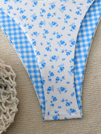 A close-up of a bikini bottom laid flat on a beige textured surface, featuring a white front panel with small blue floral print and a light blue gingham side panel.