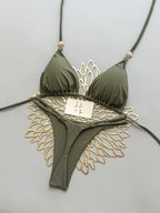 An olive green string bikini with a decorative gold metal wing-like piece behind it, laid flat on a light gray background. A small paper tag is attached near the center.