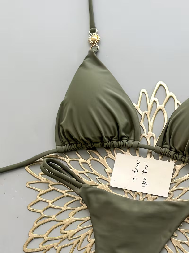 An olive green triangle bikini top and matching bottoms are laid flat on a white scalloped mat against a light gray background. A small gold sun charm hangs from the top, and a small handwritten note rests on the bikini.