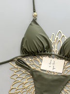 An olive green triangle bikini top and matching bottoms are laid flat on a white scalloped mat against a light gray background. A small gold sun charm hangs from the top, and a small handwritten note rests on the bikini.
