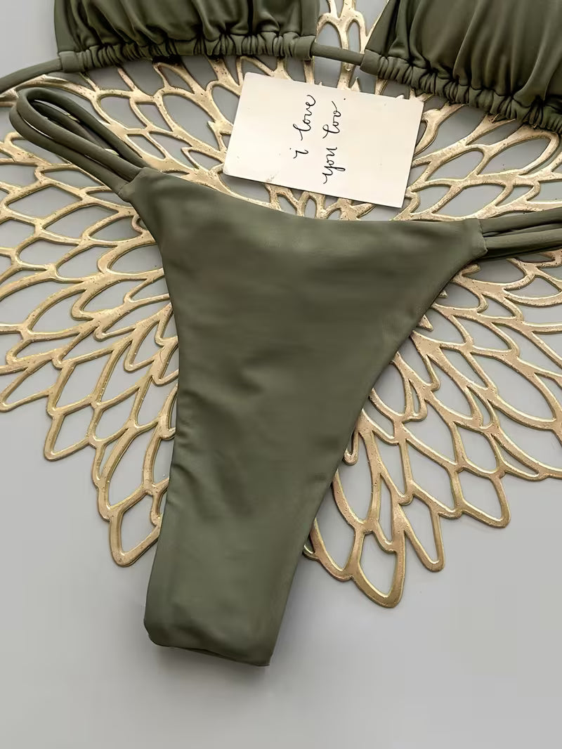 An olive green string bikini bottom laid flat on a decorative gold wire fan over a light gray background, with part of the matching string bikini top visible at the top edge and a small handwritten note attached.