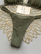 An olive green string bikini bottom laid flat on a decorative gold wire fan over a light gray background, with part of the matching string bikini top visible at the top edge and a small handwritten note attached.