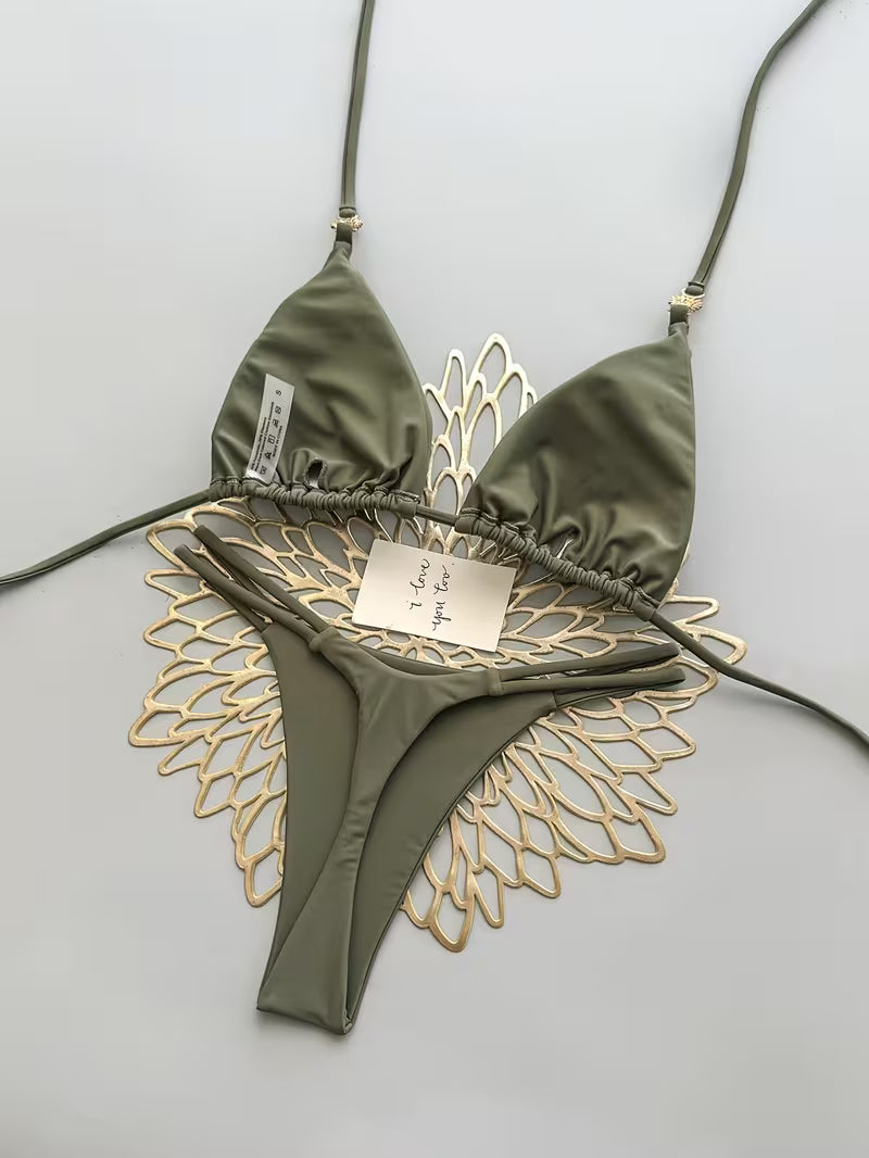 An olive green string bikini top and bottom set styled with a decorative gold metal wing-like prop on a light gray background.