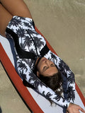 A woman lying on a beach lounge chair wearing a long-sleeve one-piece swimsuit with a black palm tree pattern on a white background.