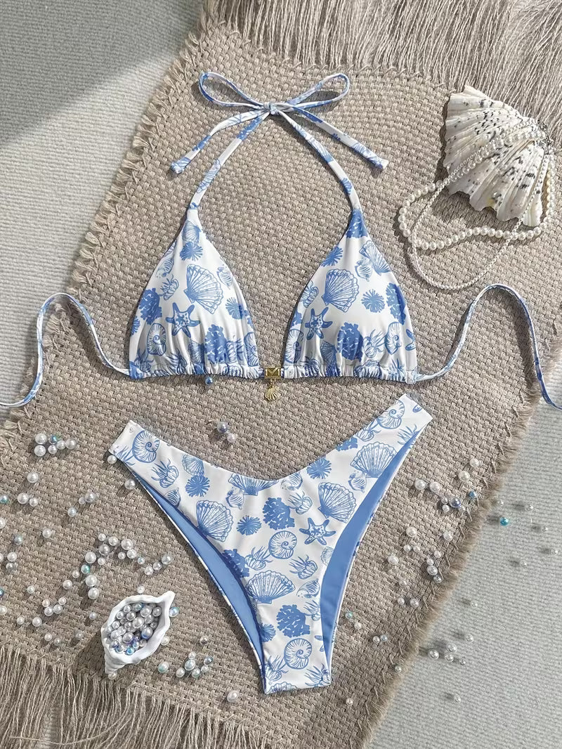 A two-piece string bikini with a blue and white seashell and sea urchin pattern, laid flat on a beige woven mat with beach-themed props including a seashell, pearls, and small blue beads.
