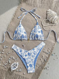 A two-piece string bikini with a blue and white seashell and sea urchin pattern, laid flat on a beige woven mat with beach-themed props including a seashell, pearls, and small blue beads.