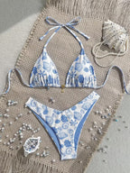 A two-piece string bikini with a blue and white seashell and sea urchin pattern, laid flat on a beige woven mat with beach-themed props including a seashell, pearls, and small blue beads.