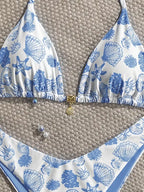 A two-piece bikini set laid flat on a woven beige surface. The triangle halter top and matching bottoms feature a white base with a blue seashell and starfish print, accented by a small gold clasp and shell charm at the center front.