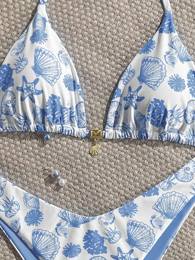 A two-piece bikini set laid flat on a woven beige surface. The triangle halter top and matching bottoms feature a white base with a blue seashell and starfish print, accented by a small gold clasp and shell charm at the center front.