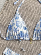 A close-up of a white bikini top with a blue seashell and starfish print, laid flat on a woven beige mat. The bikini top has thin shoulder straps, a gathered elastic hem, and a small gold shell charm at the center.