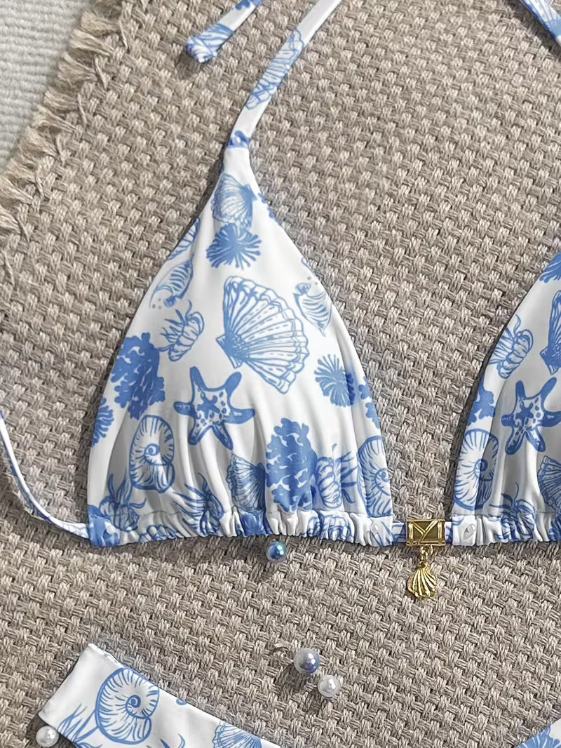 A close-up of a white bikini top with a blue seashell and starfish print, laid flat on a woven beige mat. The bikini top has thin shoulder straps, a gathered elastic hem, and a small gold shell charm at the center.