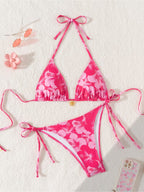 A two-piece pink floral bikini set laid flat on a textured white background, featuring a triangle halter top and matching side-tie bottoms. The bikini has a vibrant pink base with a lighter pink hibiscus flower pattern.
