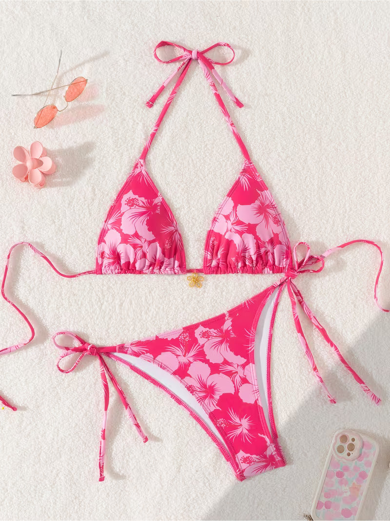 A two-piece pink floral bikini set laid flat on a textured white background, featuring a triangle halter top and matching side-tie bottoms. The bikini has a vibrant pink base with a lighter pink hibiscus flower pattern.