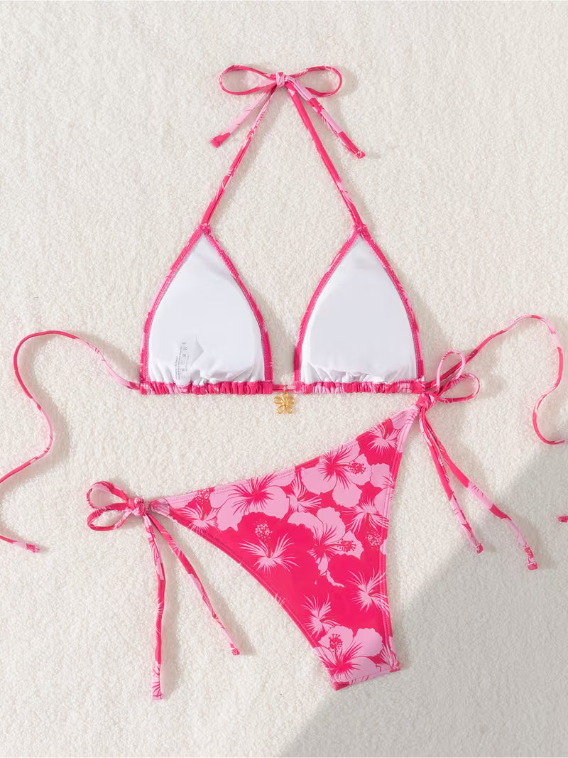 A two-piece bikini set laid flat on a textured off-white background. The top is a triangle halter style with white front panels and bright pink edges, ties, and straps.