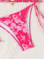 A close-up of a bikini bottom in bright pink with a large light pink and white hibiscus floral print, laid flat on a white textured fabric background.