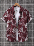 A short-sleeve button-up camp shirt with a white tropical palm leaf pattern on a deep burgundy background, styled over a white t-shirt and hanging on a wooden hanger against a gray grid wall.