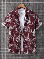 A short-sleeve button-up camp shirt with a white tropical palm leaf pattern on a deep burgundy background, styled over a white t-shirt and hanging on a wooden hanger against a gray grid wall.