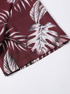 A close-up of a tropical print fabric, likely a shorts leg or shirt sleeve, featuring large monstera and palm leaves in white and light pink over a deep burgundy background.