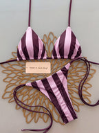 A two-piece string bikini with vertical pink and dark purple stripes, laid flat on a decorative gold metal tray over a light gray background. A small paper tag is placed in the center.