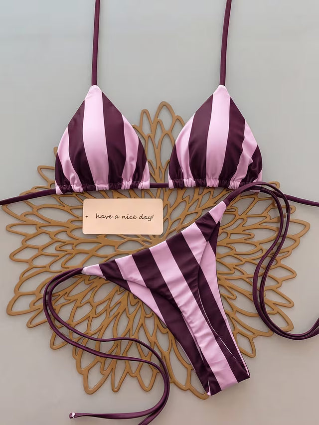 A two-piece string bikini with vertical pink and dark purple stripes, laid flat on a decorative gold metal tray over a light gray background. A small paper tag is placed in the center.