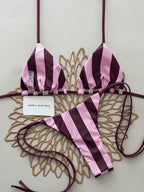 A two-piece string bikini with vertical pink and deep plum-purple stripes, laid flat on a decorative gold-colored metal tray against a light gray background.