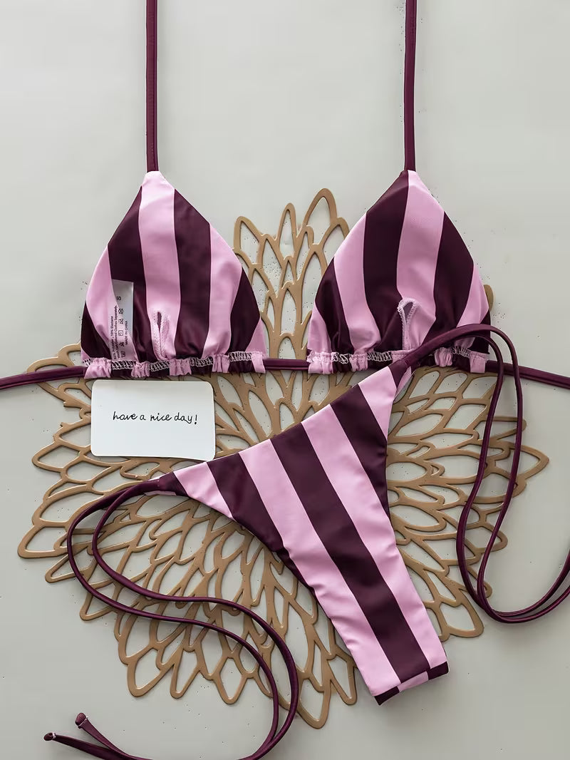 A two-piece string bikini with vertical pink and deep plum-purple stripes, laid flat on a decorative gold-colored metal tray against a light gray background.