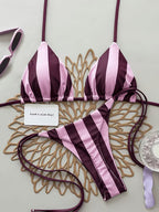 A two-piece bikini set with vertical pink and deep plum purple stripes, laid flat on a decorative gold-colored metal tray. The set includes a triangle top with thin straps and a matching low-rise string bikini bottom.