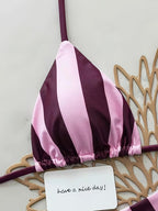 A pink and dark purple striped triangle bikini top laid flat on a light background with decorative gold paper cutouts and a small card that reads 'have a nice day!'.