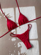 A red string bikini set with ruched details, laid flat on a textured beige wall with window shadow patterns.