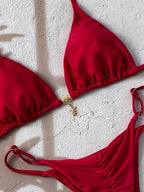 A red two-piece bikini set laid flat on a textured white surface, featuring a triangle top with a small gold clasp and matching bottoms.