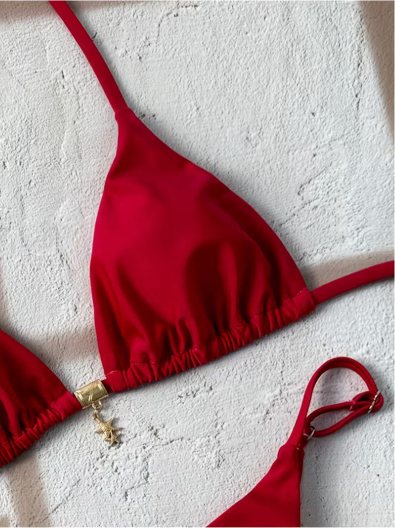 A red triangle bikini top laid flat on a textured white background, with matching red bottoms partially visible at the bottom right and a small gold clasp detail on the left side.