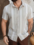 A man wearing a short-sleeve button-up shirt with vertical stripes in off-white, beige, and gray, paired with brown pants.