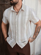 A man wearing a short-sleeve button-up shirt with vertical beige and white stripes, paired with brown trousers, standing against a rustic wooden door and white wall.