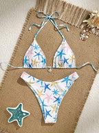 A two-piece string bikini with a white background and a colorful starfish print, laid flat on a woven beige mat with beach-themed props.
