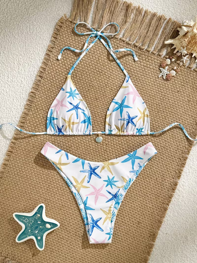 A two-piece string bikini with a white background and a colorful starfish print, laid flat on a woven beige mat with beach-themed props.