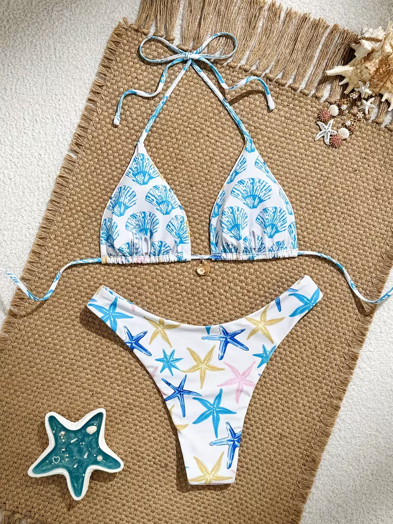 A two-piece bikini set laid flat on a woven tan mat with beach-themed props. The triangle bikini top has a white base with a blue seashell pattern, and the matching low-rise bikini bottom has a white base with blue, yellow, and pink starfish prints.