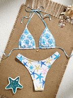 A two-piece bikini set laid flat on a woven tan mat with beach-themed props. The triangle bikini top has a white base with a blue seashell pattern, and the matching low-rise bikini bottom has a white base with blue, yellow, and pink starfish prints.