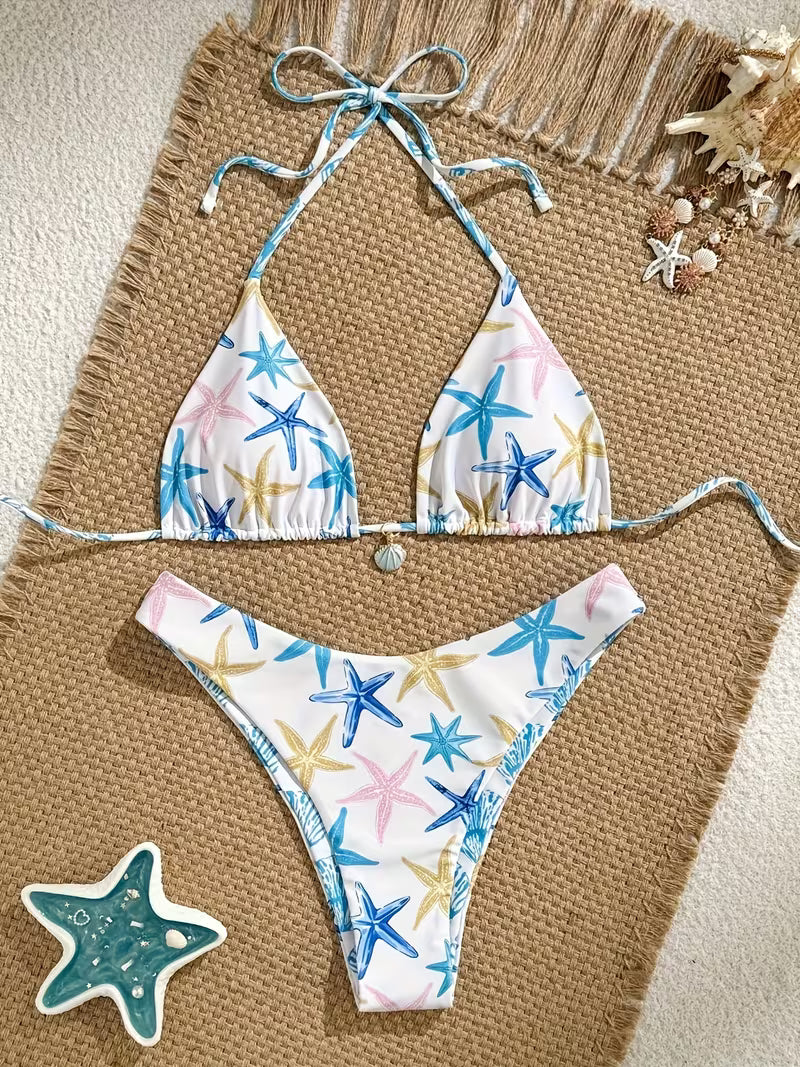 A two-piece string bikini with a white base and a colorful starfish print in blue, pink, and yellow, styled flat-lay on a brown woven mat with beach-themed props.