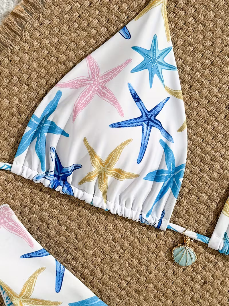 A close-up of a bikini top laid flat on a woven straw surface, featuring a white base with a colorful starfish print in blue, pink, and gold. A small seashell charm hangs from the side tie.