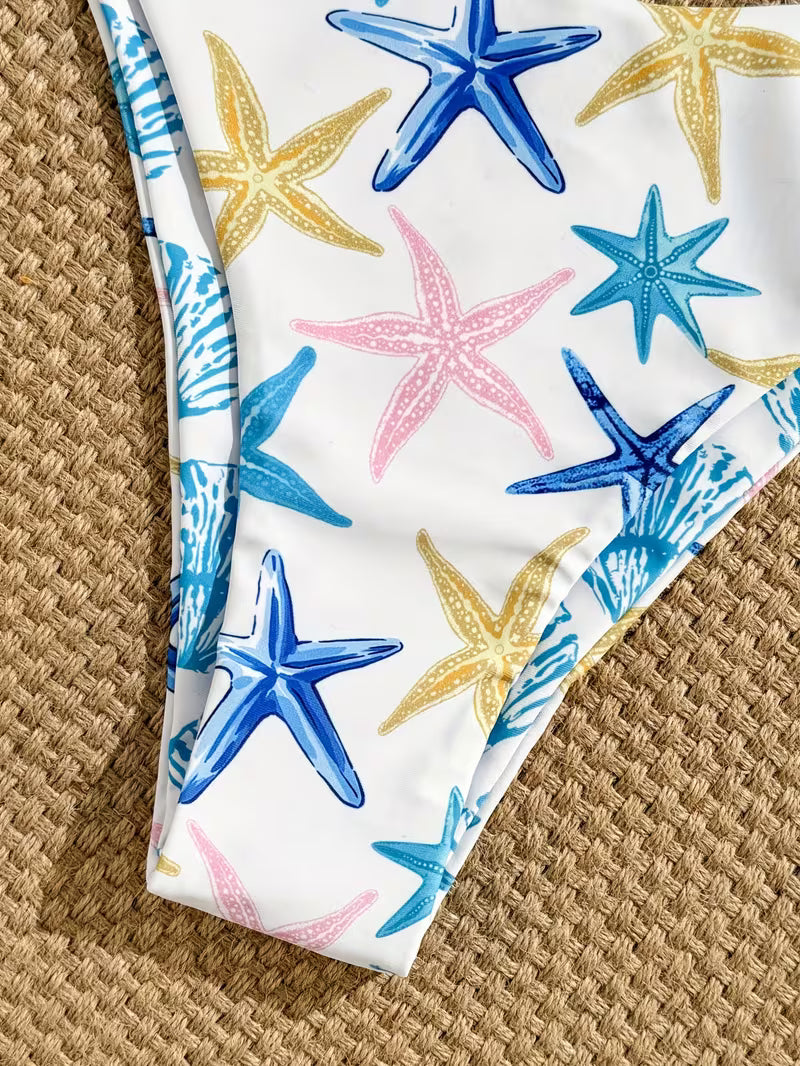 White bikini bottom with a colorful starfish print in blue, yellow, and pink, laid flat on a woven straw surface.