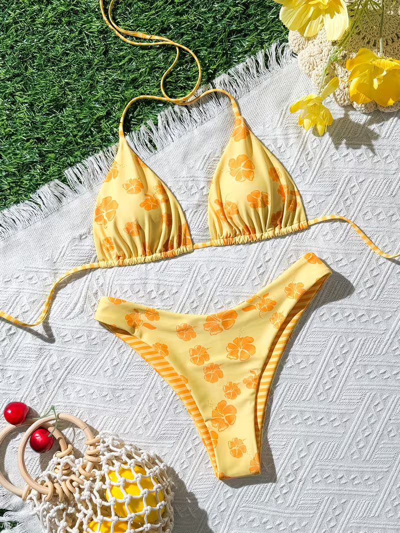 A yellow string bikini with orange floral and citrus slice patterns, laid flat on a white textured blanket with green grass and yellow flowers in the background.