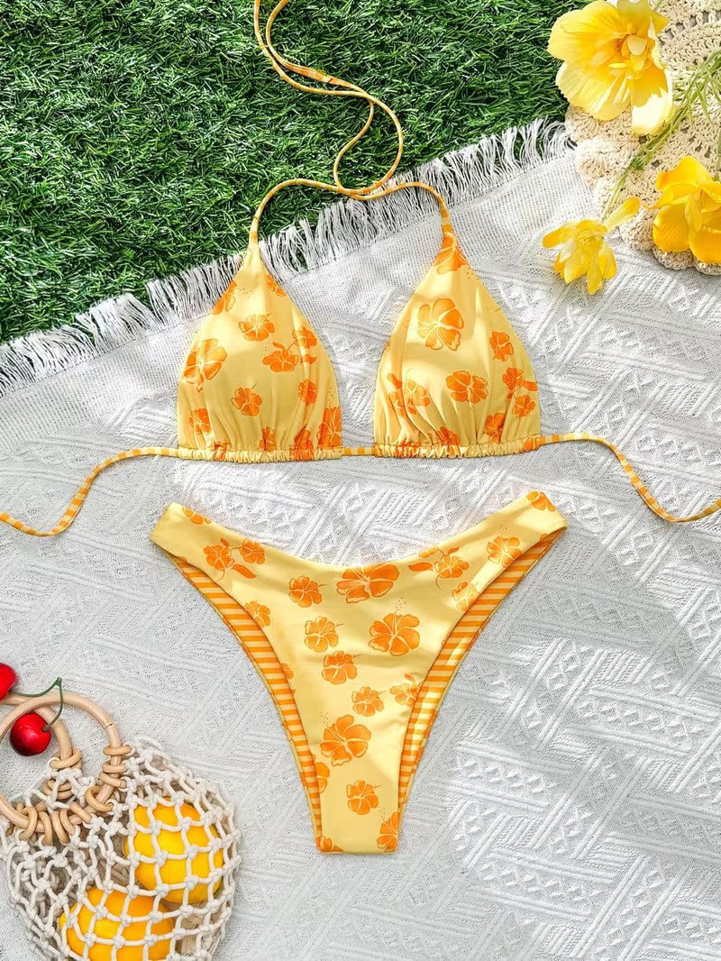 A yellow string bikini with an orange floral print and orange-and-yellow striped trim, laid flat on a white textured blanket with grass, flowers, and beach accessories around it.